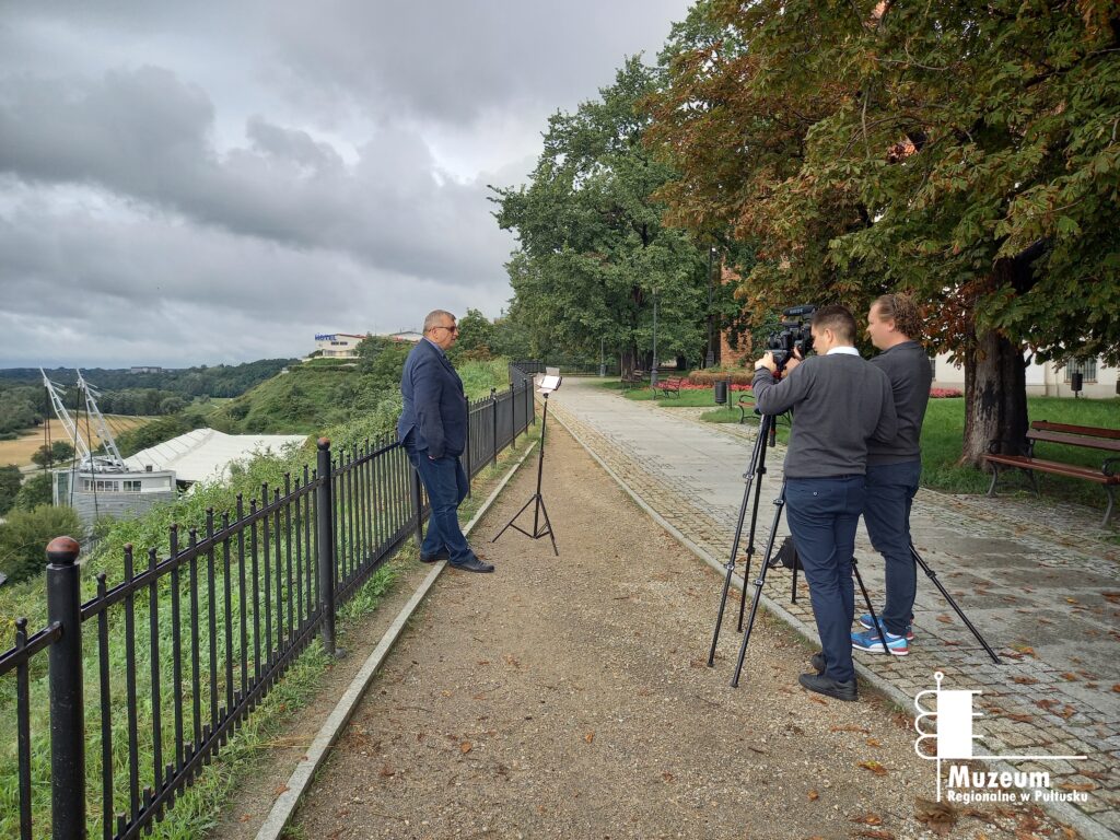 Zdjęcie wykonane na planie filmowym. Miejsce to Wzgórze Tumskie w Płocku. Po prawej stronie mężczyzna obsługujący kamerę. Przed nim stoi oparty o balustradę, za którą jest zbocze, lektor.