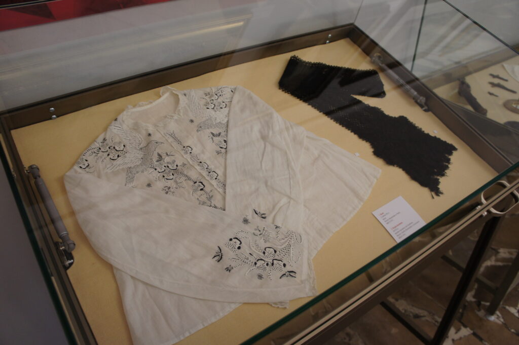 Zdjęcie wykonane w sali muzealnej. Prezentuje zawartość gabloty. Pod szklaną pokrywą na jasno-żółtym filcu ułożono białą bluzeczkę z czarnym haftem oraz czarną koronkową wstążkę. 