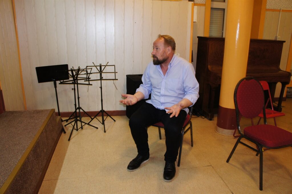 Zdjęcie wykonane podczas próby. Centralnym punktem zdjęcia jest reżyser Grzegorz Milczarczyk. Siedzi on na krzesełku. Mówi do kogoś. Gestykuluje. Ubrany w jasnoniebieską koszulę i czarne spodnie. Ma podwinięte rękawy. 