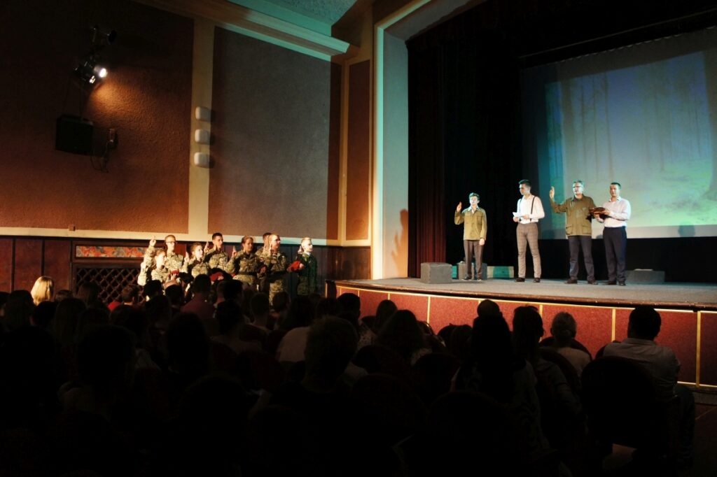 Zdjęcie wykonane podczas premiery z głębi sali. W dolnej części zdjęcia widzowie spowici mrokiem. Scena oświetlona. Na scenie czterech aktorów. Z boku sceny, po lewej stronie grupka uczniów szkoły wojskowej. Składają przysięgę.