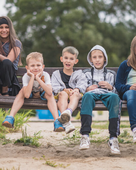 Grupa uśmiechniętych dzieci siedzi razem na dużej huśtawce ogrodowej na placu zabaw. Po lewej stronie stoi dziewczynka z lornetką, a pozostałe dzieci siedzą blisko siebie, patrząc w stronę aparatu. W tle widać drzewa i piaszczyste podłoże placu zabaw. Atmosfera radosna i wakacyjna.