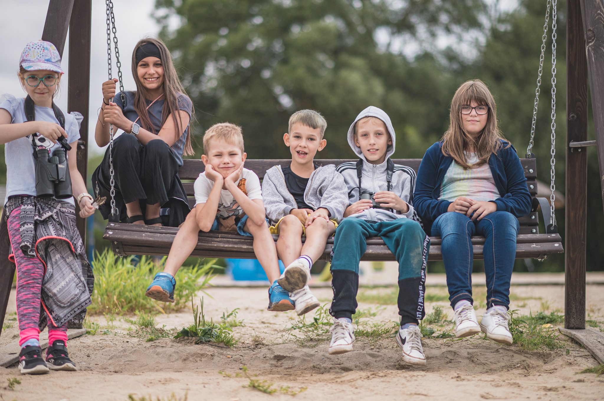 Grupa uśmiechniętych dzieci siedzi razem na dużej huśtawce ogrodowej na placu zabaw. Po lewej stronie stoi dziewczynka z lornetką, a pozostałe dzieci siedzą blisko siebie, patrząc w stronę aparatu. W tle widać drzewa i piaszczyste podłoże placu zabaw. Atmosfera radosna i wakacyjna.