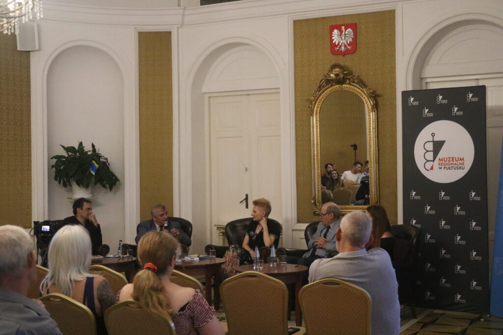 Debata w sali koncertowej Domu Polonii w Pułtusku – prelegenci siedzą w fotelach przed publicznością, mężczyzna mówi do mikrofonu. Obok stoi baner muzeum, a w lustrze w tle odbija się część sali i uczestnicy spotkania.