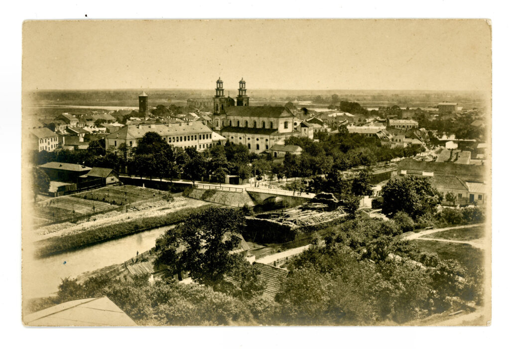 Archiwalna, czarno-biała fotografia przedstawiająca panoramę Pułtuska widzianą z podwyższonego punktu. W centrum kadru dominuje rozległy zespół dawnego kolegium jezuickiego oraz kolegiata z dwiema charakterystycznymi wieżami. Na pierwszym planie widoczna jest rzeka z kamiennym mostem, otoczona drzewami, ogrodami i zabudową gospodarczą. W tle rozciąga się gęsta zabudowa miasta oraz płaska dolina Narwi. Fotografia ma sepiowy odcień i dokumentuje historyczny wygląd Pułtuska z pierwszej połowy XX wieku.